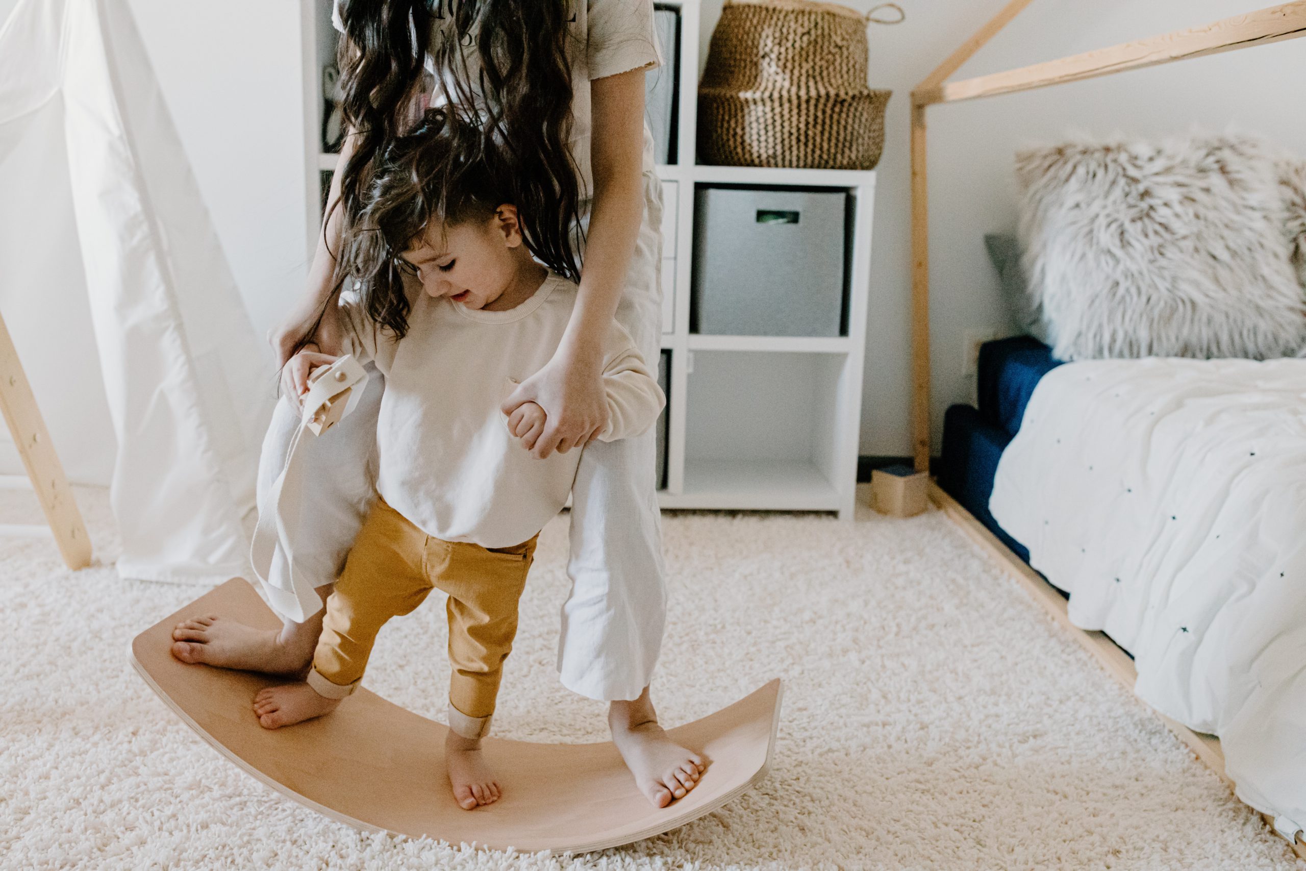 A toddler girl and her mom play on a balance board in a cute nursery.
