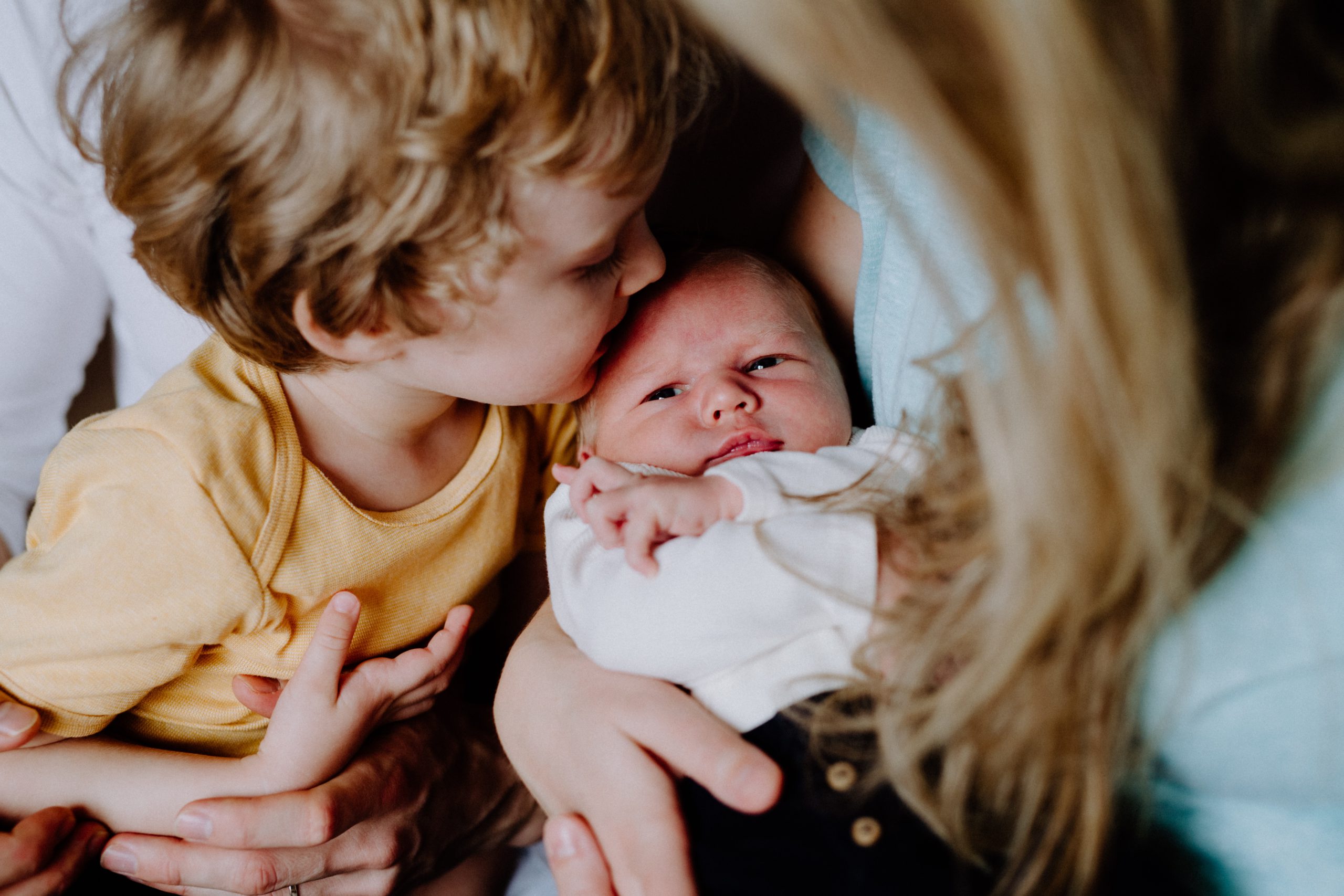 A new big brother gently kisses his newborn sister's forehad as she's snuggled in their mother's arms