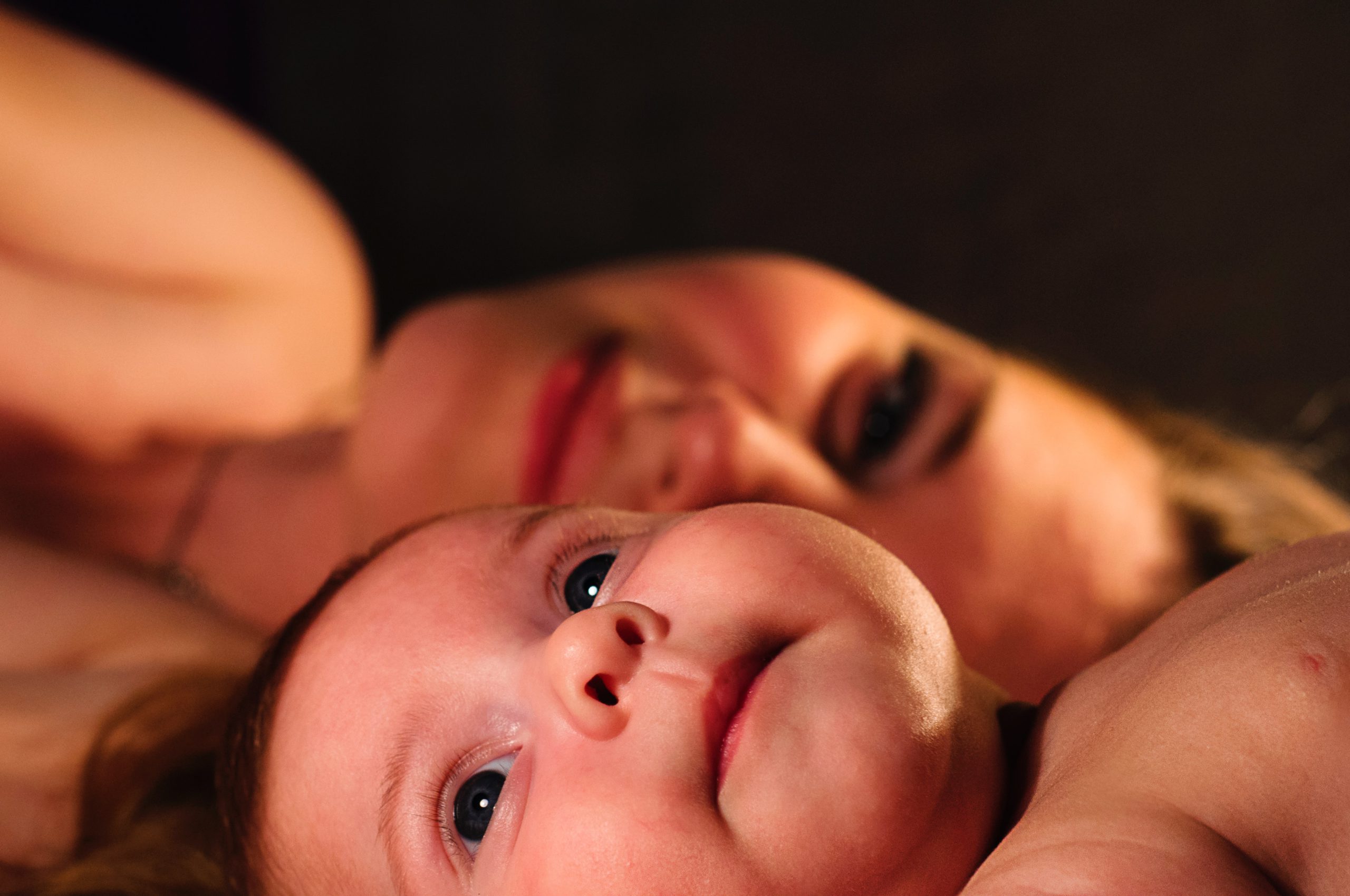 A mother and her baby laying down together. The baby looks out at something out of the frame as his mother smiles warmly at him.